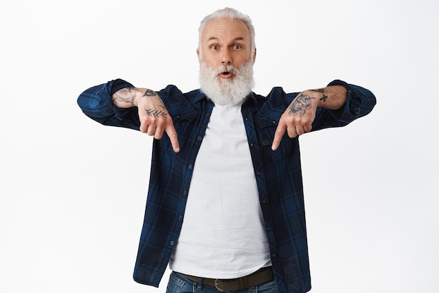 Excited senior old man with tattoos and beard gasping amazed pointing fingers down at cool advertisement showing promo offer bellow staring impressed at camera white background