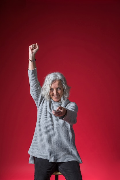 Free photo excited senior woman changing the channel with remote control cheering against red background