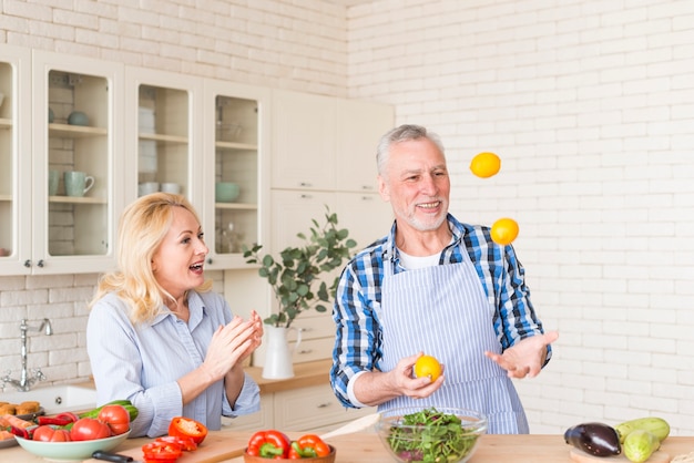 Excited senior woman clapping while her husband juggling lemons in the kitchen