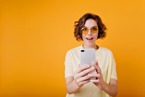 Бесплатное фото Возбужденная коротковолосая женщина держит смартфон на оранжевом пространстве