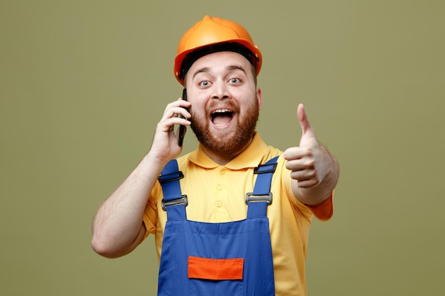 Взволнованный, показывающий палец вверх, говорит по телефону молодой строитель мужчина в униформе, изолированный на зеленом фоне