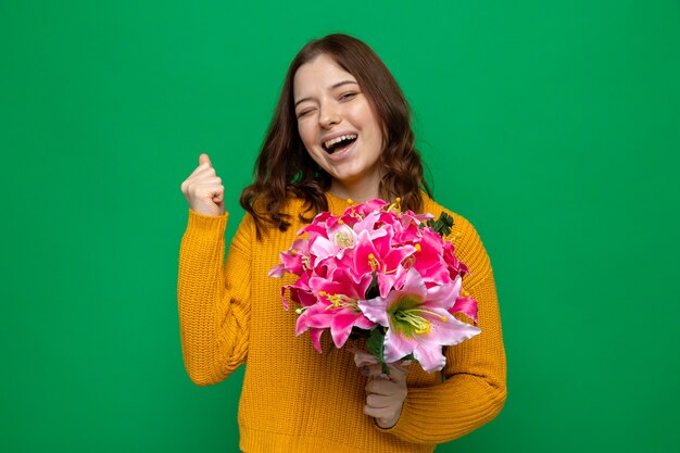Взволнованный, показывая жест да, красивая молодая девушка в счастливый женский день держит букет на зеленой стене