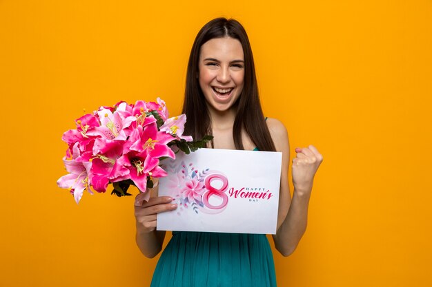 Excited showing yes gesture beautiful young girl on happy women's day holding greeting card with bouquet 