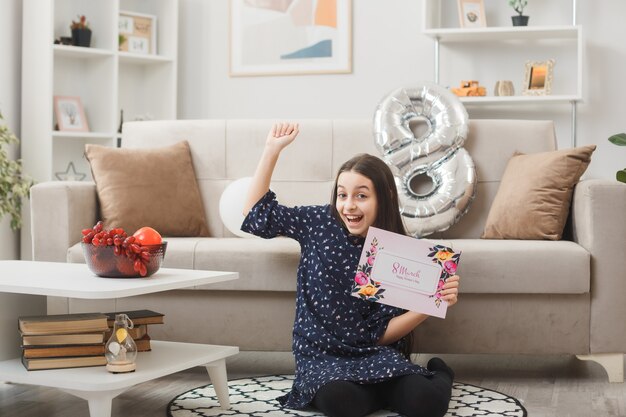 Excited showing yes gesture little girl on happy woman's day sitting on floor holding postcard in living room