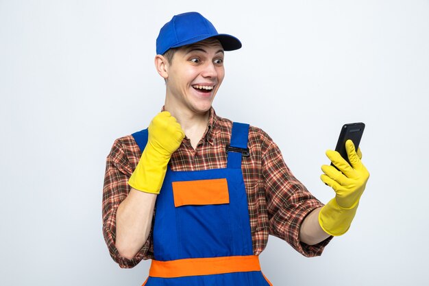 Excited showing yes gesture young cleaning guy wearing uniform and cap with gloves holding and looking at phone 