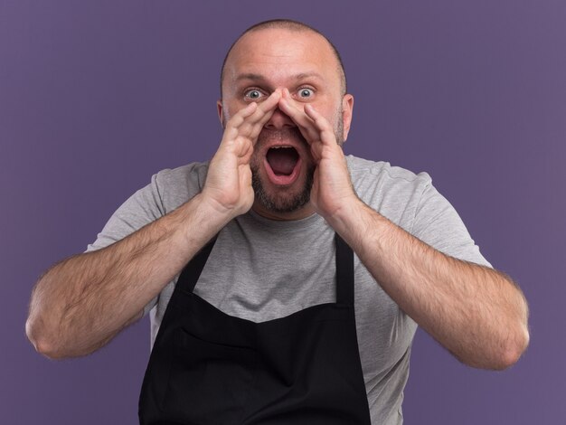 Excited slavic middle-aged male barber in uniform calling someone isolated on purple wall