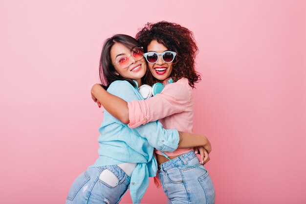 Excited slim girl with african hairstyle embracing asian female friend in trendy colorful sunglasses. Pretty european lady in jeans hugs black young woman in pink shirt.