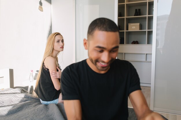 Excited smiled handsome guy is busy with tablet on bed. Upset lonely young pretty woman with long blonde hair looking at him. Having fun, relationship, at home, loneliness