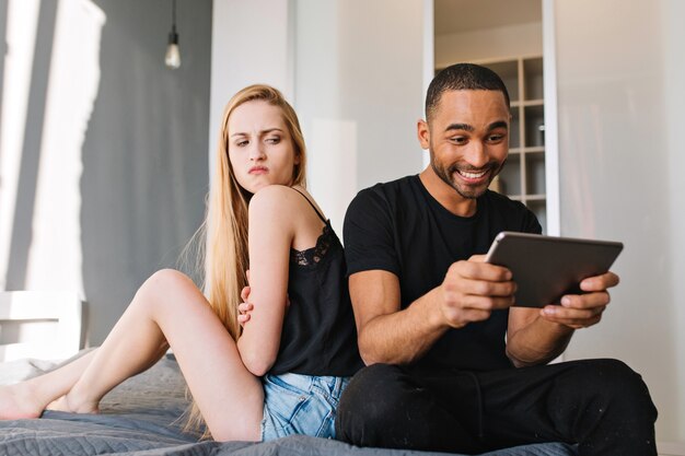 Excited smiled handsome guy surfing in internet tablet on bed near upset angry young pretty woman with long blonde hair looking at him. Having fun, relationship, at home