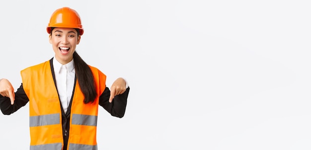 Взволнованная улыбающаяся азиатская женщина-инженер, промышленная женщина в защитном шлеме и светоотражающей куртке, указывающая пальцами вниз и делающая объявление Архитектор показывает свой проект на белом фоне