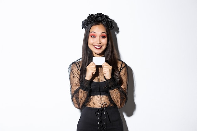 Excited smiling asian girl in black wreath and gothic witch costume showing credit card, standing over white background.