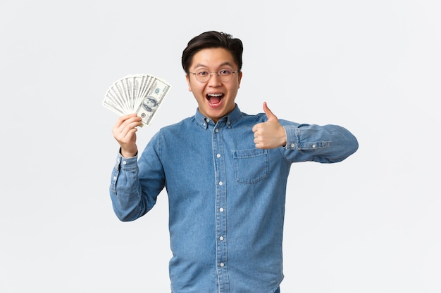 Возбужденный улыбающийся азиатский мужчина с подтяжками и очками показывает большой палец вверх и машет деньгами, получает зарплату ...