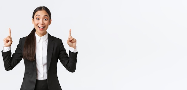 Excited smiling asian saleswoman in suit suggest great deal pointing fingers up as telling details Businesswoman making announcement and showing top banner white background