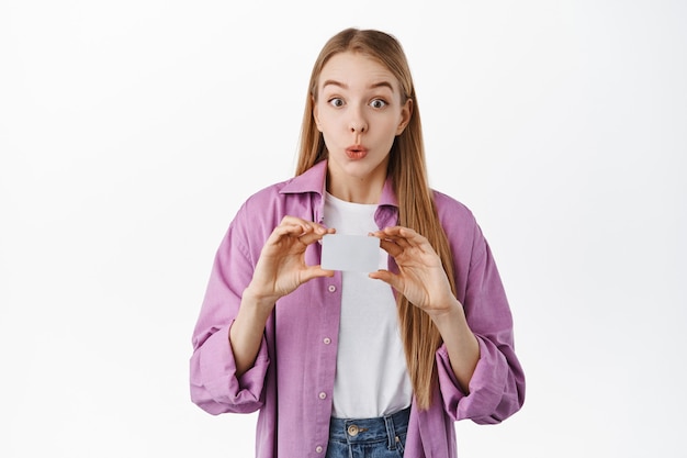 Eccitata ragazza bionda sorridente, sembra incuriosita davanti, mostrando la carta di credito della banca copyspace, in piedi sul muro bianco