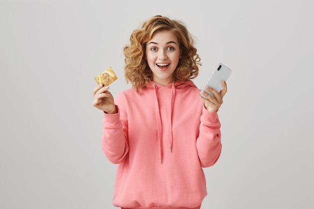 신용 카드를 보여주는 흥분 웃는 곱슬 머리 소녀, 스마트 폰을 사용하여 온라인 주문에 대한 지불