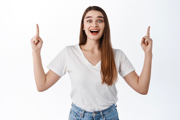 Excited smiling female student pointing fingers up, showing promo deal, advertisement banner on top, standing against white wall