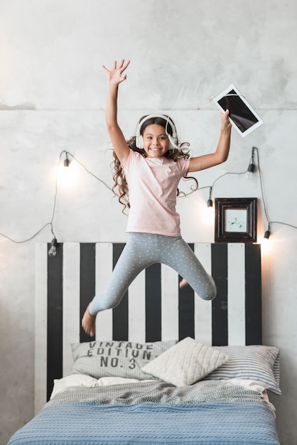 Excited smiling girl jumping on bed with headphone and digital tablet