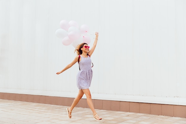 Excited smiling girl in pink sunglasses running with balloons, wearing dress and sandals