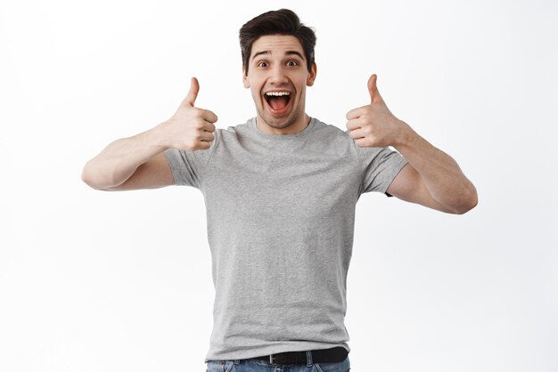 Excited smiling man shows thumbs up supportive like and praise something excellent good job well done gesture nice work standing over white background