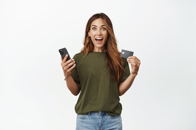 Excited smiling middle aged woman, holding smartphone, showing credit discount card