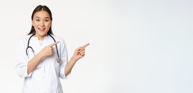 Excited smiling nurse asian doctor in medical uniform pointing right showing big promo discounts in