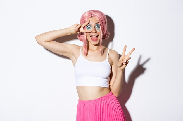 Excited smiling pretty girl in pink wig, showing peace signs and looking amazed, standing in halloween costume and pink wig.