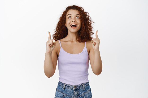 Excited smiling redhead woman, laughing and looking happy, pointing fingers up, showing promo advertisement, standing against white background