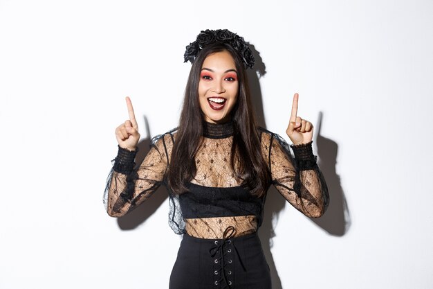 Excited smiling woman celebrating halloween in black gothic dress and wreat. Asian girl wearing witch costume and pointing fingers up, standing white background.