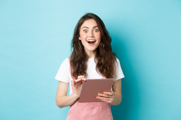 Excited smiling woman holding digital tablet, staring amazed at camera after seeing cool offer online, standing against blue background