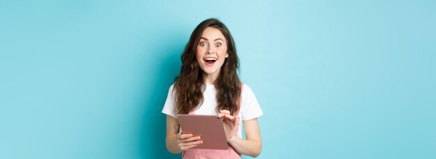 オンラインでクールなオファーを見た後、カメラに驚いて見つめるデジタルタブレットを持つ興奮した笑顔の女性