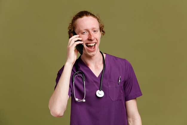 Excited speaks on the phone young male doctor wearing uniform with stethoscope isolated on green background