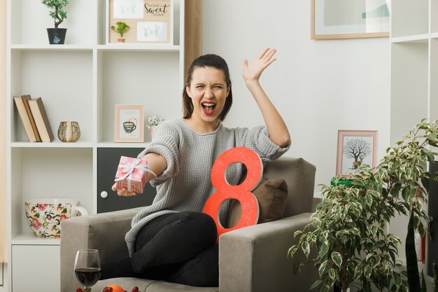 Excited spreading hand beautiful girl on happy women day holding out present at camera sitting on armchair in living room