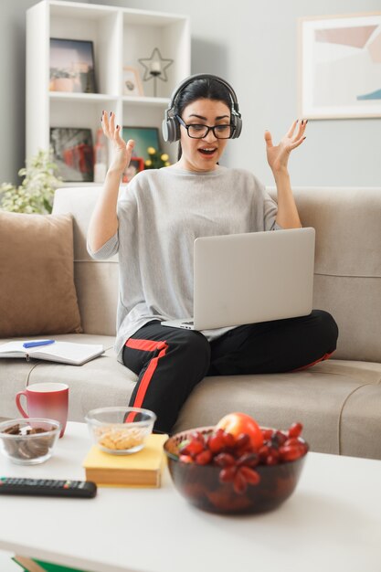 興奮して広がる手若い女の子がヘッドフォンを持って眼鏡をかけ、リビングルームのコーヒーテーブルの後ろのソファに座ってラップトップを使用しました