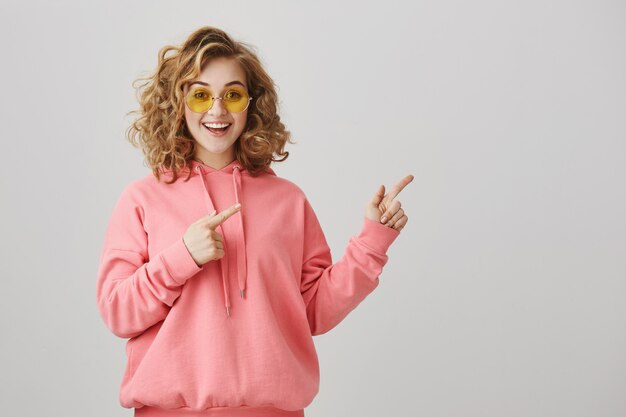 Excited stylish curly-haired girl in sunglasses pointing right, showing way