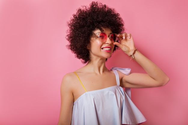 Foto gratuita eccitata ragazza alla moda con riccioli che indossa una romantica camicetta estiva e occhiali rotondi rosa che guarda da parte con il sorriso sul muro rosa