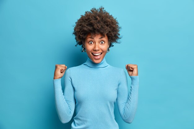 Excited successful Afro American woman clenches fists celebrates success in career says yes smiles broadly