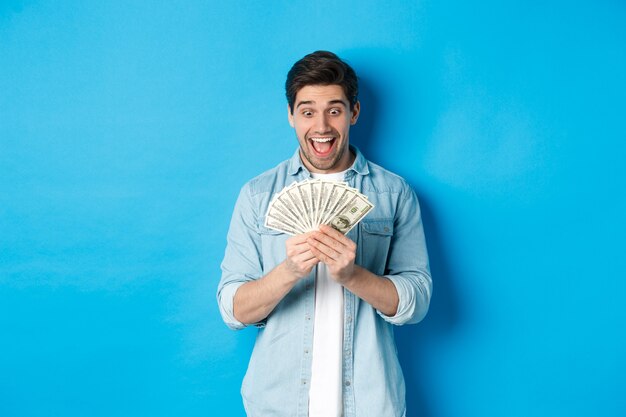 お金を数え、現金に満足しているように見え、笑顔で、青い背景の上に立って、興奮した成功した男。