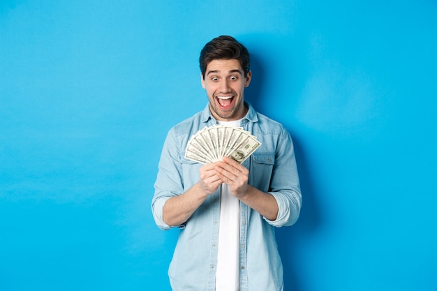 Excited successful man counting money, looking satisfied at cash and smiling, standing over blue background.