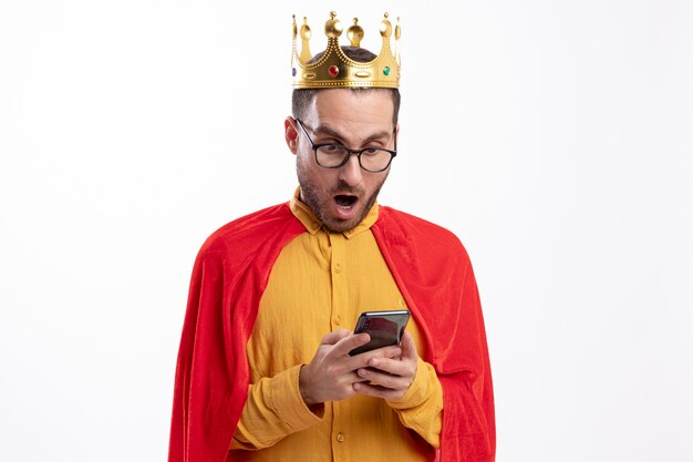 왕관과 빨간 망토와 광학 안경에 흥분된 슈퍼 히어로 남자가 보유하고 흰 벽에 고립 된 전화에서 보이는