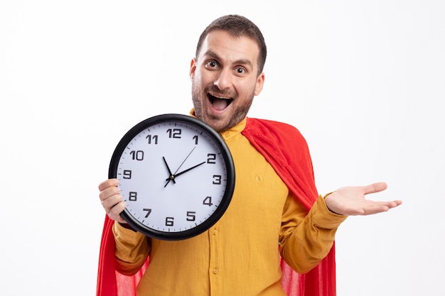 Excited superhero man with red cloak holds clock isolated on white wall