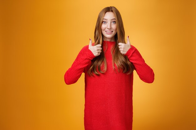 Взволнованная и поддерживающая рыжая девушка спрашивает мнение о новом теплом платье, показывая большие пальцы вверх и поднимая брови, под вопросом, ожидая мнения на оранжевом фоне