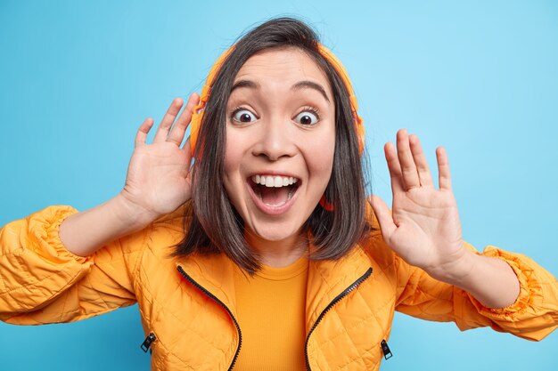 Excited suprised cheerful young Asian woman keeps mouth opened wears headphones on ears listens audio track being shocked and very happy dressed in orange jacket isolated over blue wall