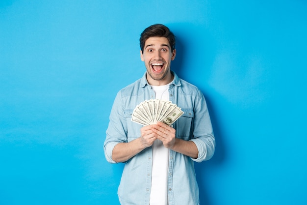 Excited and surprised attractive man, holding money prize and smiling amazed, standing over blue background