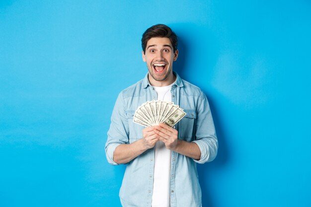 Взволнованный и удивленный привлекательный мужчина, держащий денежный приз и изумленный улыбающийся, стоящий на синем фоне