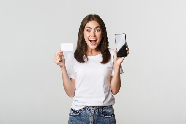 Взволнованная и удивленная милая девушка показывает на экране приложение для банковского обслуживания кредитной карты и мобильного телефона.