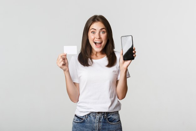 画面にクレジットカードと携帯電話のバンキングアプリを表示している興奮して驚いたかわいい女の子。