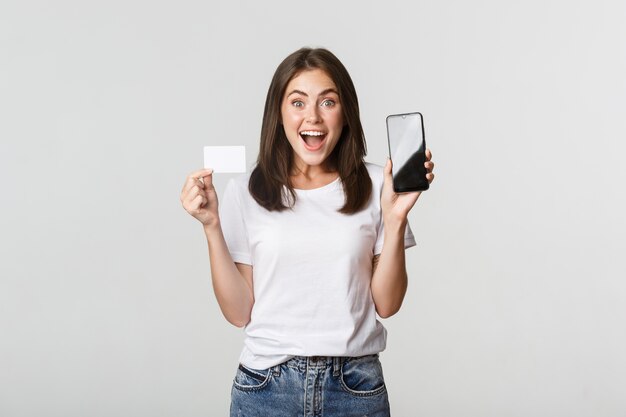 Excited and surprised cute girl showing credit card and mobile phone banking app on screen.