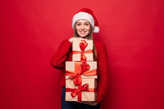 Excited surprised woman in red santa claus outfit holding stack presents isolated on the red wall