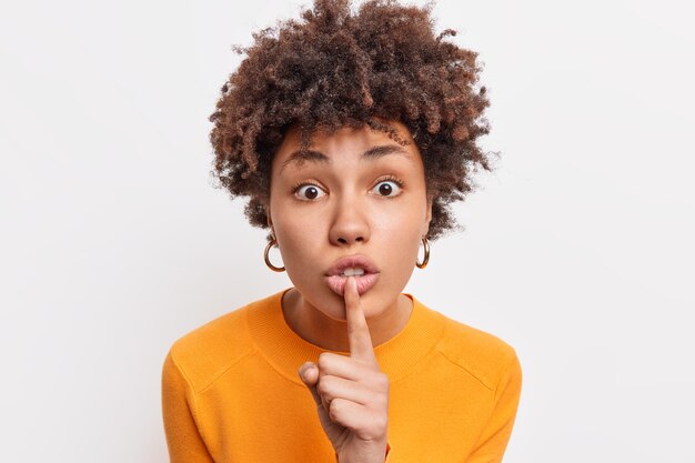 Excited surprised young Afro American female makes hush gesture gossips about something asks to stay quiet hides secret wears casual orange jumper isolated over white wall. Body language