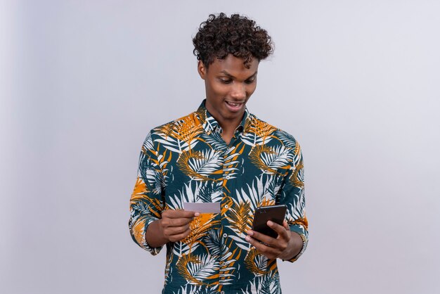 Взволнованный и удивленный молодой красивый темнокожий мужчина с вьющимися волосами в рубашке с принтом листьев смотрит в свой телефон, держа кредитную карту